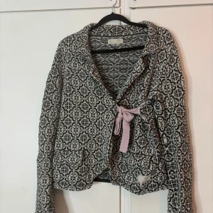 Odd Molly stickad kofta - Snygg svart och vit stickad cardigan kofta från Odd Molly med grafiskt mönster och lila knytband. Jackan har två fickor, krage och långa ärmar. Perfekt för dig som gillar unika detaljer och vill sticka ut lite extra. Där finns en tråd på innersidan av koftan (se sista bilden). Men syns ju inte eftersom det är på innersidan. Storlek 1 vilket motsvarar 38. 