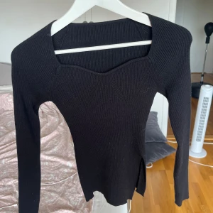 Svart ribbad stickad tröja med slits - Svart långärmad stickad tröja med ribbad struktur och snygg fyrkantig halsringning. Tröjan har en liten slits i sidan som ger en extra detalj. Perfekt för dig som gillar en stilren och enkel look med en twist.