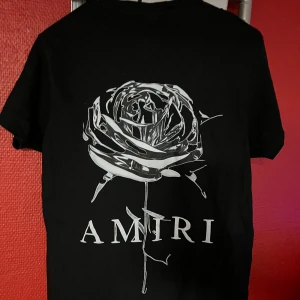 Svart Amiri t-shirt med tryck - Svart t-shirt från Amiri med coolt tryck av en silvrig ros och logga på ryggen samt färgstänk och Amiri-tryck på bröstet. Klassisk passform och rund hals. Perfekt för dig som vill sticka ut med streetstyle-vibe.