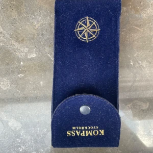 Blått fodral från Kompass Stockholm - Mörkblått fodral i mjukt material från Kompass Stockholm, perfekt för att förvara dina glasögon. Fodralet har en guldfärgad kompassros på framsidan och stängs med en knapp. Snygg och stilren design som skyddar dina glasögon.