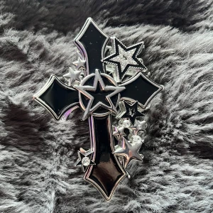 Stor svart hårklämma med kors och stjärnor - Cool hårklämma i form av ett svart kors med silverdetaljer och flera stjärnor, vissa med glittrande stenar. Klämman har en edgy vibe och är perfekt för att ge din frisyr en unik touch. Metall och plast i silver och svart, med blingiga detaljer. Köpt från hottopic i USA FL