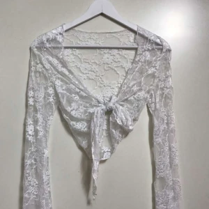 Vit spetsig omlottopp med knyt - Superfin vit långärmad topp i spets med omlott och knyt framtill. Transparent och lätt material med blommigt spetsmönster, perfekt att styla över ett linne eller bralette.  ONE SIZE som passar om man har storlek S, M och även L.