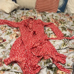 Röd blommig omlottklänning Amelie & Me - Supersöt röd omlottklänning från Amelie & Me med vitt blommönster. Klänningen har långa ärmar, volangdetaljer och knytband i midjan. Perfekt för dig som gillar en feminin och lekfull stil.