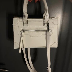 Vit handväska med dragkedja - Stilren vit handväska i skinnimitation med både handtag och avtagbar axelrem. Väskan har silverfärgade detaljer, en praktisk ytterficka med dragkedja och ett rymligt innerfack. Perfekt för att bära dina viktigaste grejer med snygg look.