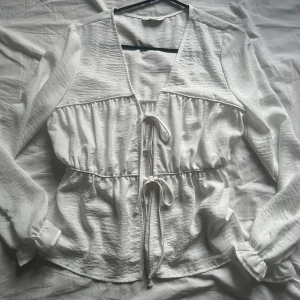 Vit blus - Supersöt vit blus med v-ringning och två knyt framtill. Långa ärmar med resår vid handlederna och lätt rynkad struktur som ger en luftig känsla. Perfekt för dig som gillar romantiska och stilrena plagg.