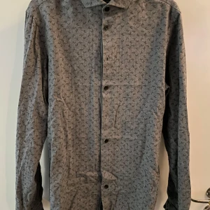 Grå mönstrad skjorta från Casual Friday - Snygg grå skjorta från Casual Friday i slim fit-modell. Skjortan har ett diskret svart mönster över hela tyget, klassisk krage och svarta knappar framtill. Perfekt för dig som gillar stilrena detaljer och en smalare passform.