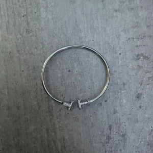 Silverfärgat armband med T-detaljer - Stilrent armband i silverfärg med öppen design och två T-formade ändar, varav ena sidan är dekorerad med små glittrande stenar. Perfekt för dig som gillar minimalistiska smycken med en touch av bling.