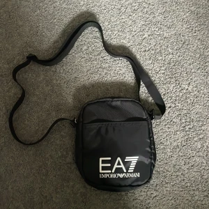 Svart axelväska EA7 Emporio Armani - Snygg svart axelväska från EA7 Emporio Armani med justerbar rem och stort vitt logotryck på framsidan. Väskan har dragkedja med metall-detalj och är tillverkad i slitstark polyester. Perfekt för att bära dina viktigaste grejer med stil.