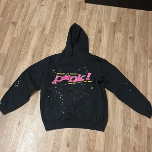 Svart hoodie ”P*nk!” från Sp5der, storlek L. - Cool svart hoodie från Sp5der i 100% bomull med färgglad print på bröstet och stjärndetaljer på ärmarna. Stor luva och relaxed fit, perfekt för dig som gillar streetstyle. Unik design med spindelnätsmönster och tryck i rosa, gult och blått.