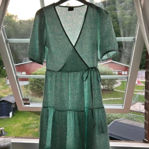 Ljusblå omlottklänning med vita blommor - Superslt miniklänning med småblommigt mönster och omlottmodell. Klänningen har puffiga kortärmar, knyt i midjan och v-ringning.