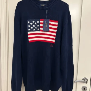Mörkblå stickad tröja Polo Ralph Lauren - Mörkblå stickad tröja från Polo Ralph Lauren med amerikansk flagga framtill och RL-broderi. Klassisk rund halsringning och långa ärmar. Perfekt för dig som gillar preppy stil och vill sticka ut med en ikonisk detalj.