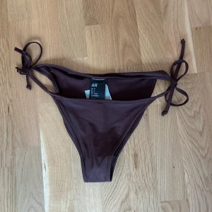 HELT NY bikiniunderdel - Ny, helt oanvänd snygg brun bikiniunderdel från H&M med knyt i sidorna. Jag råkade köpa i fel storlek, har inte ens provat den och lapparna är kvar. Modellen är enkel och stilren. Perfekt för strandhäng eller poolparty!