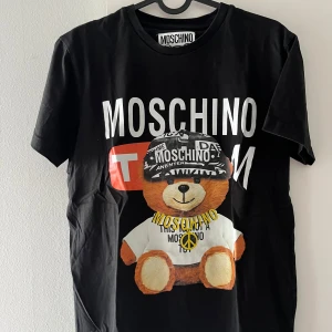 Svart Moschino t-shirt med björntryck - Svart t-shirt från Moschino med stort tryck av en nallebjörn i keps och t-shirt framtill. Tydlig Moschino-logga både på bröstet och på björnens keps. Klassisk rund hals och korta ärmar. Perfekt statement-plagg för dig som gillar streetstyle.