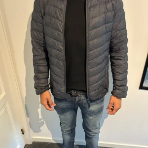 Mörkgrå north bend pufferjacka herr - Snygg mörkgrå pufferjacka med quiltad design och dragkedja framtill. Jackan har lång ärm, diskret patch på ena ärmen och två fickor med dragkedja. Perfekt för kyliga dagar och enkel att matcha med jeans eller hoodie. 