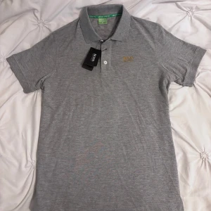 Hugo Boss T-shirt  - Snygg grå pikétröja från Hugo Boss med klassisk krage och två knappar framtill. Tröjan har broderad gul logga på bröstet och är tillverkad i mjuk bomull. Perfekt för en clean och stilren look.