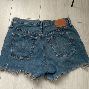 Jeansshorts från Levi's - Säljer ett par klassiska Levi's 501 jeansshorts i blå denim. De har en knappgylf och fransiga kanter för en avslappnad look. Perfekta för sommardagar!