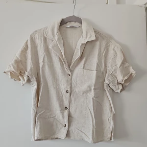 Beige kortärmad skjorta från Stradivarius - Säljer en stilren beige skjorta från Stradivarius. Skjortan har korta ärmar och knappar framtill. Perfekt för en avslappnad look.