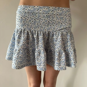 Blå blommig kortkjol - Söt blå kortkjol med blommigt mönster och volangdetaljer. Perfekt för en somrig look. Kjolen har en bekväm passform och är lätt att matcha med olika toppar.