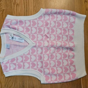Rosa och vit stickad väst från Lindex - Säljer en söt rosa och vit stickad väst från Lindex med hjärtmönster. Västen har en v-ringad design och ribbade kanter. Perfekt för att lägga till en gullig touch till vilken outfit som helst. 💖