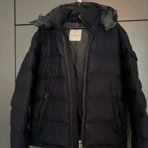 Svart dunjacka från Moncler - Säljer en stilren svart dunjacka från Moncler med dragkedja och huva. Jackan har långa ärmar och en broderad logotyp på ärmen. Perfekt för kyliga dagar. En liten defekt vid handleden därför sänks priset.