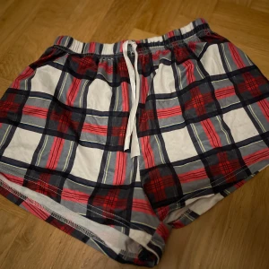 Rutiga pyjamas shorts med snörning - Säljer ett par bekväma shorts med rutig design i rött, blått och vitt. De har en praktisk snörning i midjan för justerbar passform. Perfekta för en avslappnad stil.