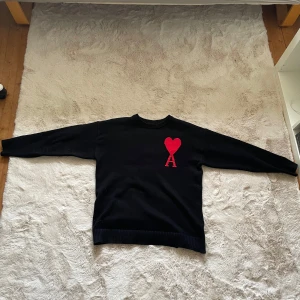 Svart tröja med rött hjärtmönster från Ami - Säljer en svart tröja från Ami med ett stort rött hjärta och bokstaven A på framsidan. Tröjan har långa ärmar och är tillverkad i ett mjukt material. Perfekt för en stilren look.