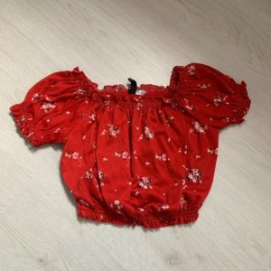 Röd blommig offshoulder topp - Säljer en söt röd offshoulder topp med blommigt mönster. Toppen har korta puffärmar och elastiska kanter för en bekväm passform. Perfekt för sommarens varma dagar!
