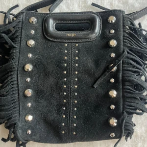 Svart mockaväska med fransar från Maje - Snygg svart axelväska i mocka från Maje med coola fransdetaljer och nitar. Väskan har ett praktiskt handtag och en stilren design som passar perfekt för att ge din outfit en edgy touch. Perfekt för när man ska iväg men inte vill ha stor väska då den ändå rymmer mycket🖤strlk mini! Nypris ca 2500