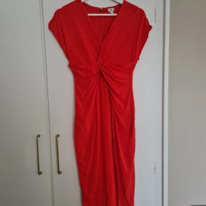 Röd klänning från River Island - Snygg röd klänning från River Island med v-ringad design och knytning i midjan. Klänningen har korta ärmar och en elegant drapering framtill. Klänningen har också en slits framtill. Aldrig använd, endast testad! 