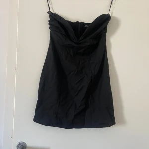 Svart axelbandslös klänning - Snygg svart axelbandslös klänning. Perfekt för en elegant kvällslook. Klänningen har en draperad detalj framtill som ger en stilren touch.