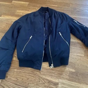 Mörkblå bomberjacka  - Snygg mörkblå bomberjacka  med dragkedjor framtill och på ärmen. Jackan har en klassisk bomberstil med ribbade muddar vid ärmslut och nederkant. Perfekt för en cool och avslappnad look. Köpte för 250