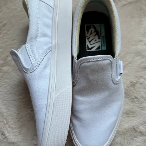Vita slip-on sneakers från Vans - Säljer ett par klassiska vita slip-on sneakers från Vans. Skorna har en enkel och stilren design med elastiska paneler för enkel på- och avtagning. Perfekta för studentflaket! Storlek 36.