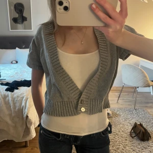 Grå kofta från In Extenso - Säljer en stilren grå kofta från In Extenso. Den har en djup v-ringning och korta ärmar, perfekt för lager-på-lager. Koftan är croppad med knappar framtill och ribbade detaljer vid ärmslut och nederkant.