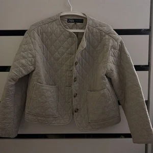 Beige quiltad jacka från Zara - Säljer en beige quiltad jacka från Zara storlek S  med knappar framtill och två stora fickor. Jackan har en rund halsringning och långa ärmar. Perfekt för en stilren look. Har en liten defekt framtill men är inte jätte anmärkningsbar.