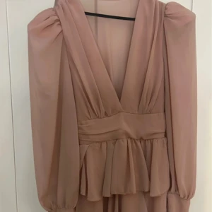 Beige långklänning med v-ringning - Elegant beige långklänning med v-ringning och långa ärmar. Klänningen har en smickrande passform med rynkade detaljer och ett band i midjan. Perfekt för en stilfull kväll.