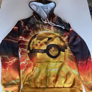 Färgglad Pikachu-hoodie - Säljer en färgglad hoodie med Pikachu-motiv. Tröjan har en elektrisk design med blixtar i gult, orange och svart. Den har en stor ficka framtill och en justerbar huva med vita snören.
