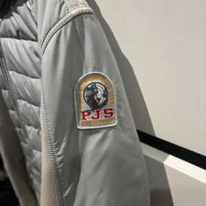 Ljusblå jacka från Parajumpers - Säljer en ljusblå jacka från Parajumpers med huva och dragkedja. Jackan har quiltade detaljer och en patch på ärmen. Perfekt för kyligare dagar.