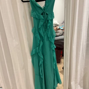 Grön långklänning med volanger - Elegant grön långklänning med volanger och knytning i ryggen. Perfekt för sommarens festligheter. Klänningen har en v-ringad design och är ärmlös, vilket ger en luftig och stilren look.