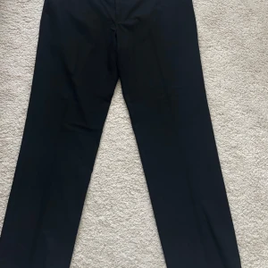 Svarta kostymbyxor från Dressmann - Snygga svarta kostymbyxor från Dressmann med en klassisk rak passform. Perfekta för formella tillfällen eller en stilren vardagslook. Byxorna har en enkel design utan extra detaljer.