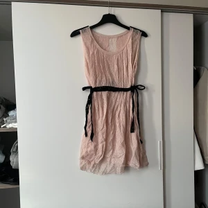 Ljusrosa spetsklänning med svart knytband - Säljer denna super söta ljusrosa klänning i spets med ett svart knytband i midjan. Klänningen är ärmlös och har en luftig passform men även lite genomskinlig, perfekt för varma dagar. Den har en fin struktur och faller vackert.💞💞