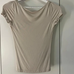 Beige kortärmad topp med öppen rygg - Snygg beige topp med korta ärmar och en djup öppen rygg. Perfekt för en stilren och luftig look. Tillverkad i ett mjukt och bekvämt material. Använd en gång