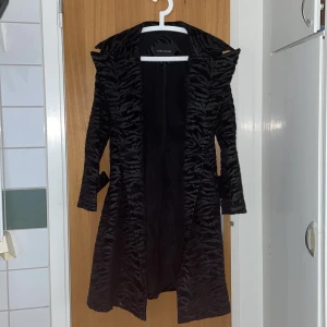 Svart mönstrad kappa från Zara Basic - Snygg svart kappa från Zara Basic med ett subtilt/Zebra mönster. Den har långa ärmar och ett bälte i midjan för en elegant look. Perfekt för att ge din outfit en stilfull touch.