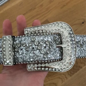 Glittrigt bälte med strass - Säljer ett glittrigt bälte med strassdetaljer och en stor, dekorerad spänne. Bältet är i en iögonfallande silverfärg och passar perfekt för att ge en extra touch till din outfit.