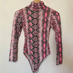 Rosa ormmönstrad bodysuit från Boohoo - Säljer en cool rosa ormmönstrad bodysuit från Boohoo. Den har en polokrage och långa ärmar. Materialet är mjukt och stretchigt för en bekväm passform.