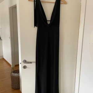 Svart balklänning med öppen rygg - Elegant svart långklänning med djup v-ringning och öppen rygg. Klänningen har en smickrande passform och är perfekt för festliga tillfällen. Den är ärmlös och har en stilren design som ger en sofistikerad look. Nypris: 599