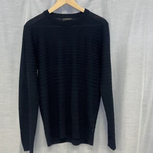 Svart tröja från Emporio Armani - Snygg svart tröja från Emporio Armani med diskret randigt mönster. Tröjan har långa ärmar och en rund halsringning, perfekt för en stilren look. Passar bra till både jeans och chinos.