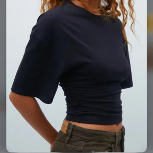 Svart kortärmad topp med rynkdetalj - Snygg svart kortärmad topp med rynkdetalj i sidan. Perfekt för en stilren look. Tillverkad i mjukt material som ger en bekväm passform. Passar utmärkt till både jeans och kjol.