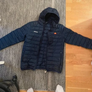 Mörkblå pufferjacka från Ellesse - Säljer en mörkblå pufferjacka från Ellesse med huva och dragkedja. Jackan har långa ärmar och är quiltad för extra värme. Perfekt för kyligare dagar. Köpt för 1000kr så denna är 600kr billigare 