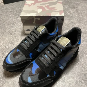 Valentino Rockrunner sneakers i blått och svart - Snygga Valentino Rockrunner sneakers med ett coolt blått och svart camouflagemönster. Skorna har snörning och en rund tå, vilket ger en bekväm passform. Perfekta för att ge din outfit en unik touch. Helt oanvända