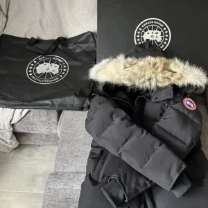 Svart dunjacka från Canada Goose - Säljer en svart dunjacka från Canada Goose med en lyxig pälsfodrad huva. Jackan har en klassisk design med en broderad logotyp på ärmen och knappar framtill. Perfekt för kalla vinterdagar. 